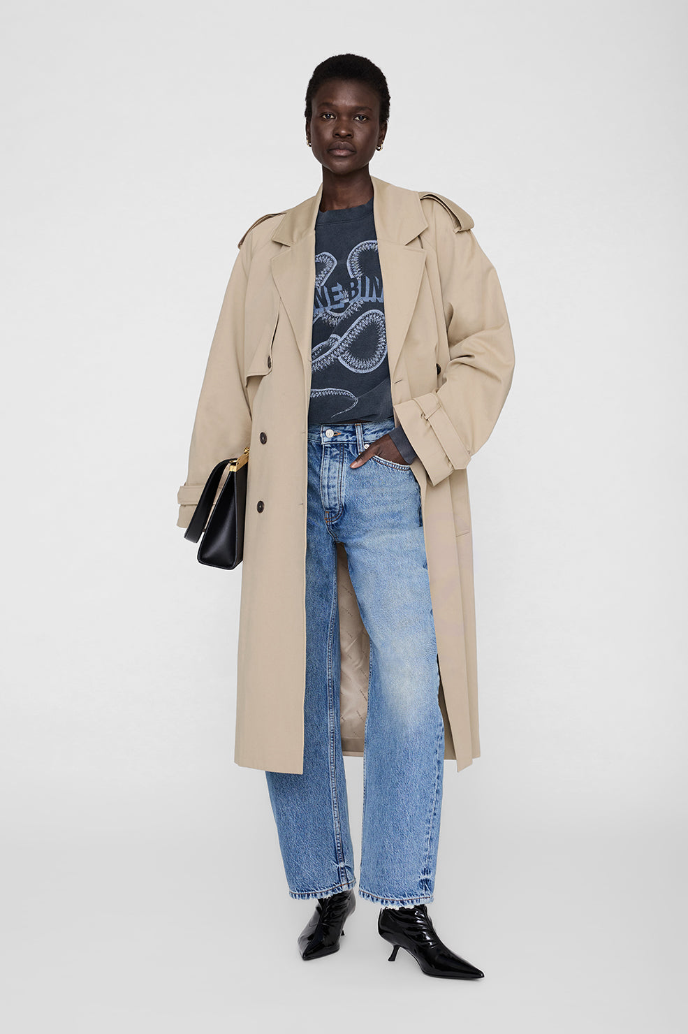 Person wearing a beige trench coat, graphic t-shirt, blue jeans, and black shoes on a white background